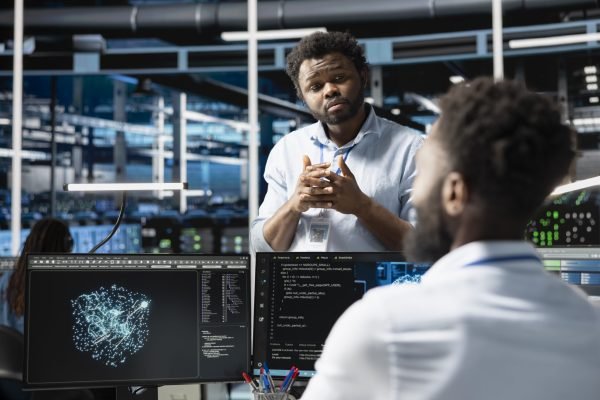 Server farm manager supervising engineer looking at data analysis graph to reduce failures. Data center team leader overseeing worker looking at AI visualization dashboard to pinpoint gear glitches